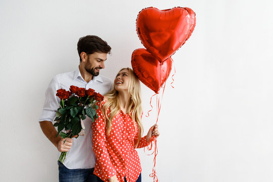 Couple. Love. Valentine's day. Emotions. Man is giving heart-shaped balloons to his woman, both smiling; on a white background