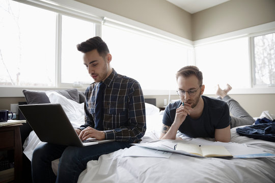 Gay male couple working from home, using laptop and reading on bed