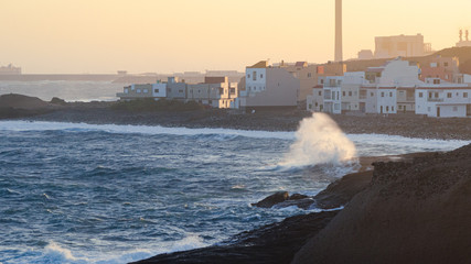 Waves hitting the rocky shore with houses and an industrial harbour with large chimney in the background, with copy space