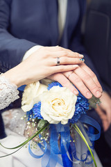 Obraz na płótnie Canvas Hands of newlyweds with wedding rings, on a bouquet of white roses with classic blue ribbons. The concept of love and marriage. Accessories for the bride and groom.
