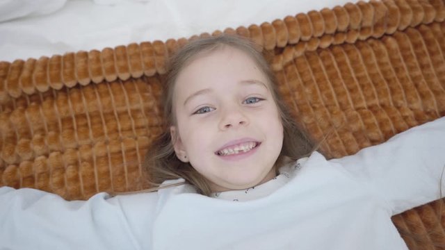 Top view of cute Caucasian child enjoying weekends at home. Cheerful little girl falling back on bed and laughing, camera moving around kid's face. Joy, childhood, leisure.