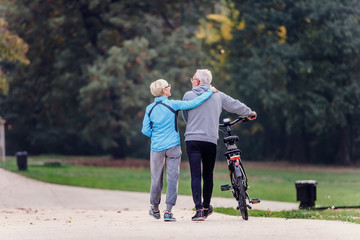 Cheerful active senior couple with bicycle walking through park together. Perfect activities for elderly people.