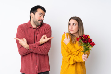 Couple in Valentine Day holding flowers over isolated background pointing to the laterals having doubts