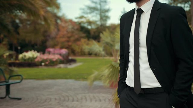 Young slender brunette businessman with beard in black suit thin tie is walking along road to his goal around beautiful trees bushes palm trees. Puts his hands in pocket