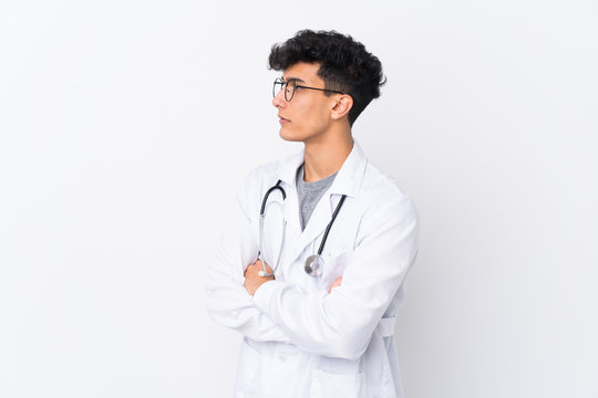 Young Argentinian man over isolated white background wearing a doctor gown and with arms crossed looking side