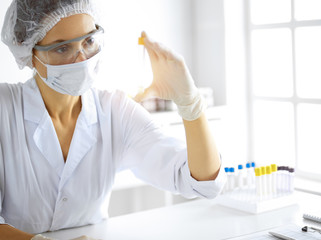 Professional female scientist in protective eyeglasses researching tube with reagents in sunny...