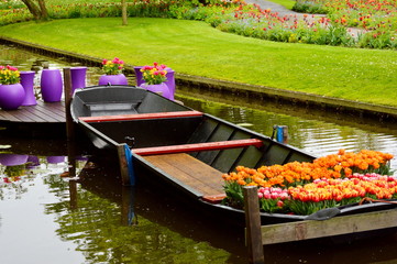 Obraz premium Tulip flowers on wooden boat in venice. Floral tulip market floating on river in green colorful park, violet vase pots. Dinghy craft boat tulip shop in venice city garden canal as tourism background