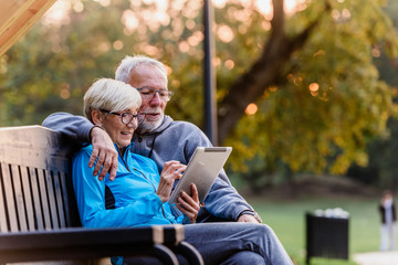 Smiling senior active couple sitting on the bench looking at tablet computer. Using modern technology by elderly. - obrazy, fototapety, plakaty