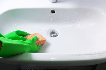 Hands in green rubber gloves clean sink in domestic bathroom yellow sponge and liquid foam...