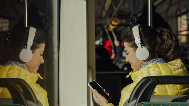 Young woman smiling while standing in train, tram or bus. Happy female passenger listening to music on a smartphone in public transportation.