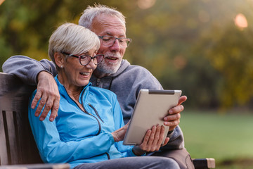 Smiling senior active couple sitting on the bench looking at tablet computer. Using modern...