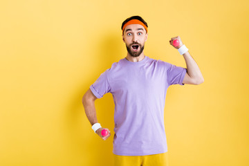 surprised bearded man looking at camera while holding small dumbbells on yellow