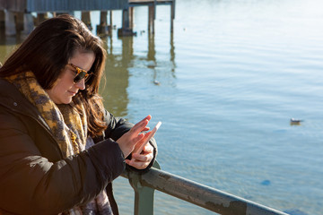 Young lady is sitting on a bench of a table at the pier of a lake. She is using her smartphone, also she is wearing sunglasses and winter clothes.