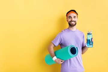 positive bearded man holding sports bottle and fitness mat on yellow