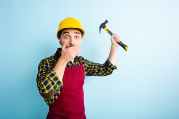 shocked workman holding hammer and covering mouth on blue