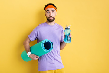 bearded man holding sports bottle and fitness mat on yellow