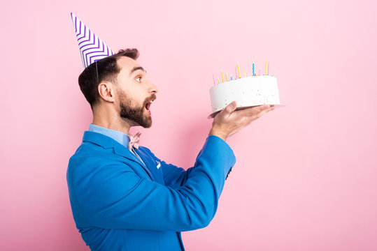 side view of emotional businessman in party cap looking at birthday cake on pink