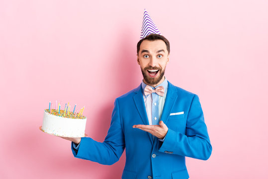excited businessman in party cap pointing with hand at birthday cake on pink