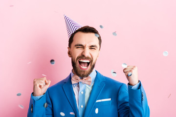 excited businessman in party cap celebrating triumph near falling confetti on pink