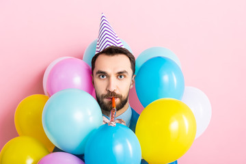 bearded man in party cap with party blower in mouth near balloons on pink