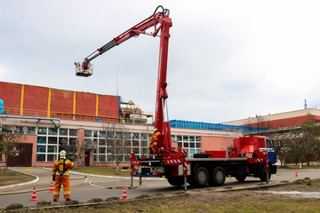 Firefighter rescuers at work in fireproof suits came to extinguish a fire in a fire truck and pull out a ladder cradle lift at a large industrial plant with pipes and equipment.
