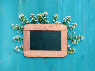 Blackboard with white cutter flower scattered around on light blue wooden background. Flowers composition. Flat lay. Copy space on blackboard.
