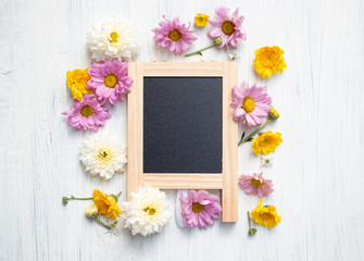 Little chalkboard sign with wooden frame and many flowers on white wooden background.  Flowers composition. Flat lay. Copy space on chalkboard sign.