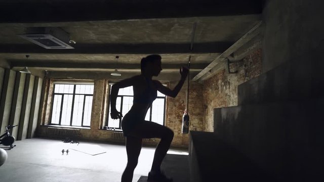 Girl in silhouette doing exercises, stretching and warming in gym. Healthy lifestyle. Side view, camera moving with sports woman running up on stone stairs in dark loft interior. 