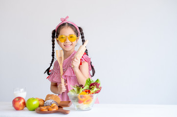 Cute Asian girl shows kitchenware with various types of fruit and vegetables and stand in front of white wall background.