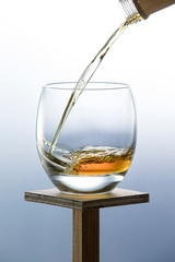 Whiskey pouring from the neck of a bottle into a glass on a wooden stand