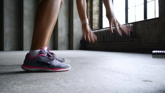 Young athletic woman warming up before workout in the gym. Cropped shot flexibility girl does morning stretch and aerobic burpee exercises on floor in dark loft interior opposite big window