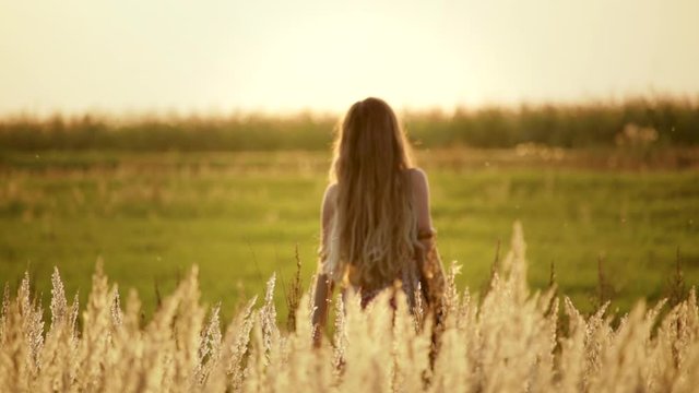 Lifestyle: portrait of a brown-haired woman in a floral print dress with a SLR camera on a shoulder strap in a field with high dry grass at sunset. Slow motion of a hipster girl posing