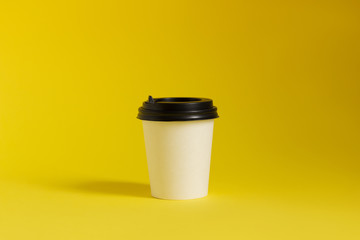 White coffee paper cup with black cap on yellow background. Copy space for brand. Coffee to go cup. 