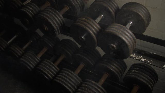 Pan Across Of Weights Lined Up On Weight Rack In Industrial Gym Old Style Old Fashioned Under Ground Gym. Paint Work Scratched Off Smoke Passing Over