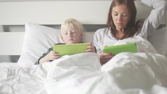 Mom and son lie in bed in the morning and check social networks on tablets. Awakening with gadgets