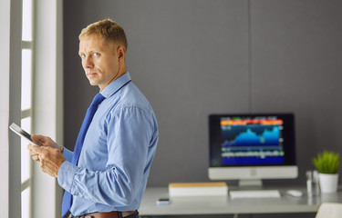 Portrait of smiling happy young entrepreneur standing at his office table and working on tablet computer