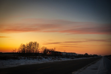 An industrial freight train illuminated by headlights travelling at sunset along a deserted highway in a prairie winter landscape