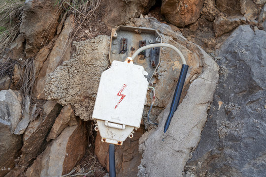 Outdoor electrical cable, leading to old junction box with lightning drown on the cover. Set in big stones and concrete.