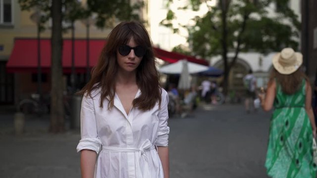 Gimbal shot of woman dressed in white dress and wearing black sunglasses, walking alone on the road on background on Munich pedestrian zone in summer day