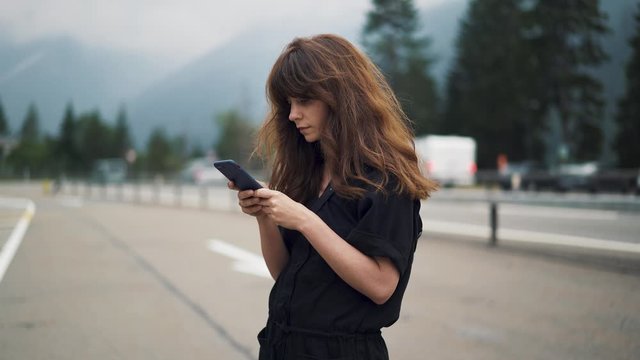 Gimbal shot of woman typing with the phone typing texting standing on blurred background of highway road car lanes and mountains with forest
