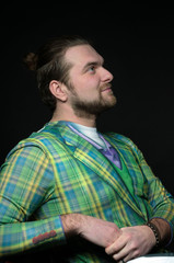 side view of handsome bearded man with hair bun in green hipster blazer leaning with clenched hands on chair and looking away isolated on black