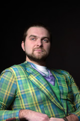 handsome bearded man in green hipster blazer leaning with clenched hands on chair and looking away isolated on black