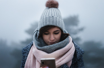 Front portrait of young and pretty natural girl texting and looking on the phone in winter time. Female model - woman in winter outfit (coat, cap, scarf) is browsing web on smartphone in misty forest.