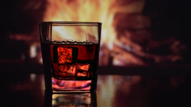 Glass whiskey, scotch, bourbon or cognac with ice stands on a table against background of a fireplace with a flame. Alcoholic drink concept. Fire background. Close up. 4K