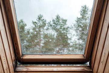 An ajar roof window in the attic, with raindrops dripping down the glass and distorting the image.