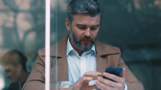Rotation view of handsome middle aged man with grey hair and beard uses phone drinks coffee at coffee shop texing message working successful people daily routine relaxing communicating slow motion
