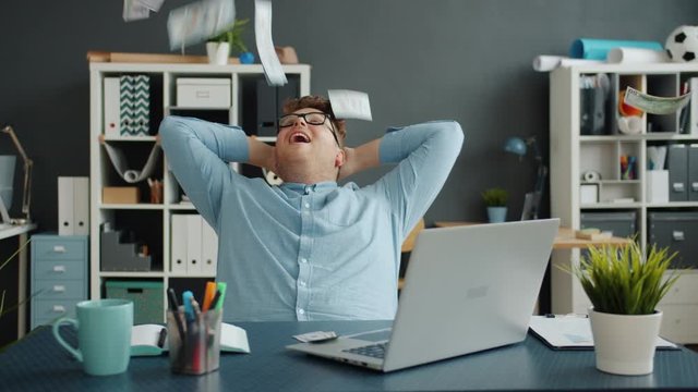 Slow motion of lots of money falling on handsome guy in office sitting at desk alone enjoying wealth. Happiness, rich youth and financial luck concept.