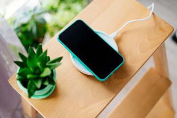 Smartphone in mint silicone case is charged from a wireless charger. The mobile phone is charged on a wooden nightstand or shelf.