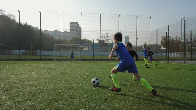 Young forward taking pass, masterfully bypassing back player and scoring goal during football match between children's teams. Active preteen footballer showing good skill while playing soccer outdoors