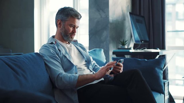 Handsome caucasian mature man sitting on the sofa at home and using phone watching video or contents serfing internet technology modern apartment social media browsing isolated